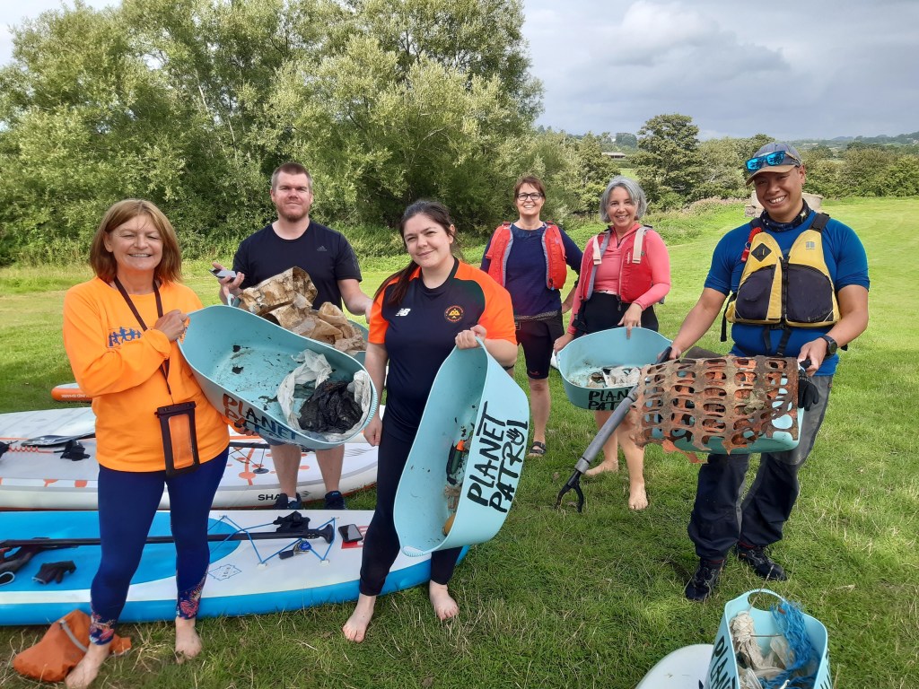 A group of volunteers holding containers filled with litter collected from the river, standing on a grassy area with paddleboards in the background.