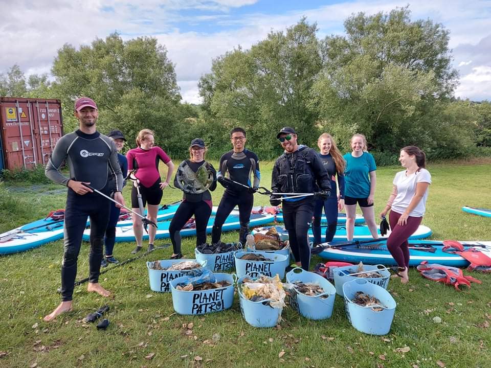A group of volunteers standing together, surrounded by kayaks and holding litter-picking tools, with containers filled with collected river debris labeled 'Planet Patrol' in the foreground.
